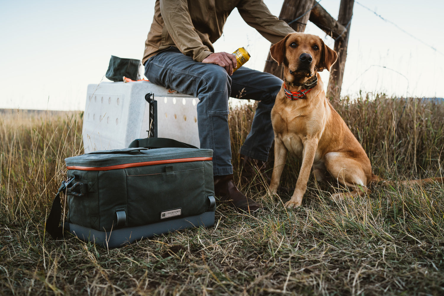 Person sitting with a dog in a field next to a cooler and a bottle.
