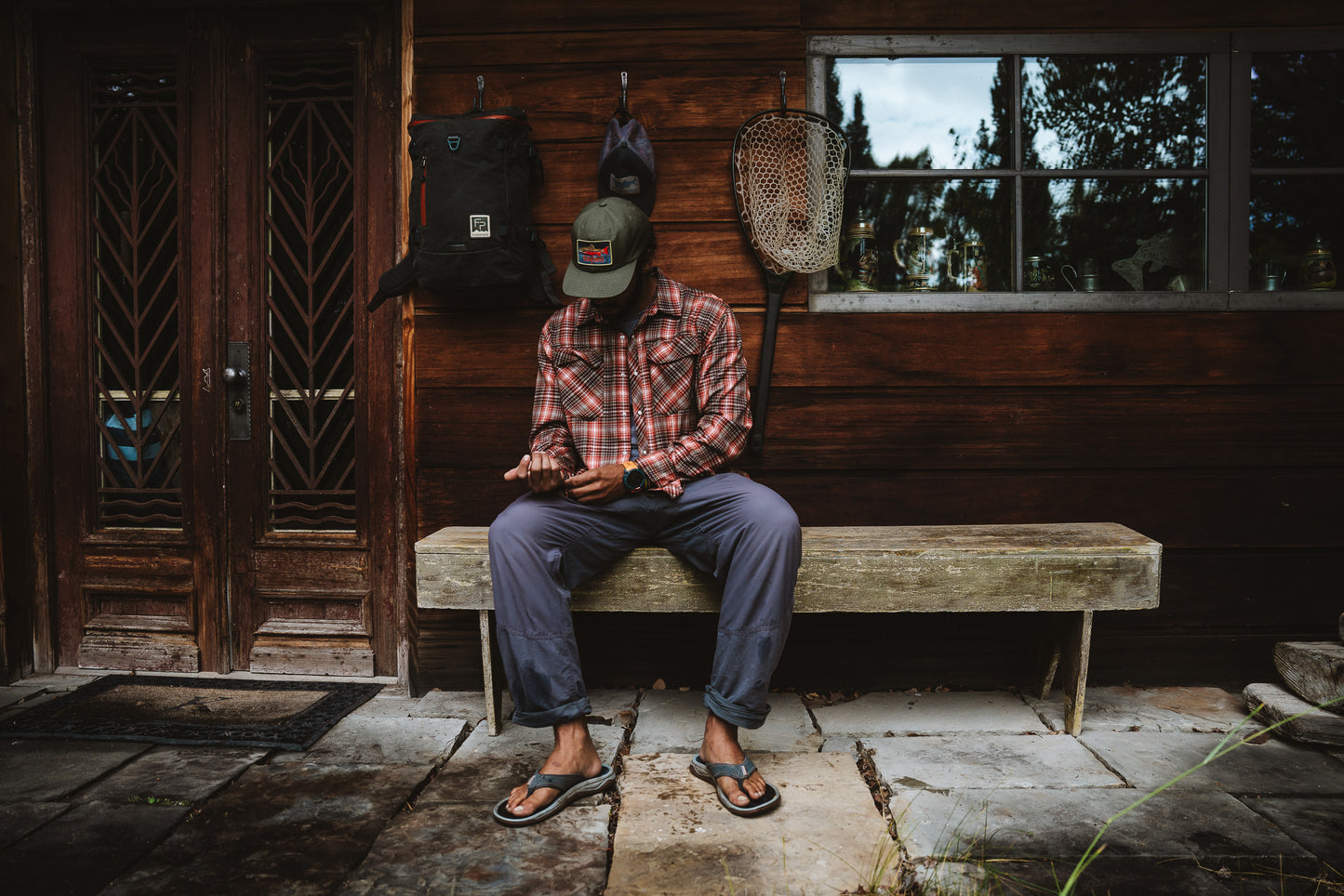 Person sitting on a wooden bench outside a cabin with outdoor gear.